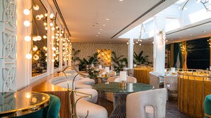 A modern interior with a round glass table surrounded by white cushioned chairs, a bar area with a wooden counter, and a large window providing natural light. - The Delborough Lagos (Lagos)