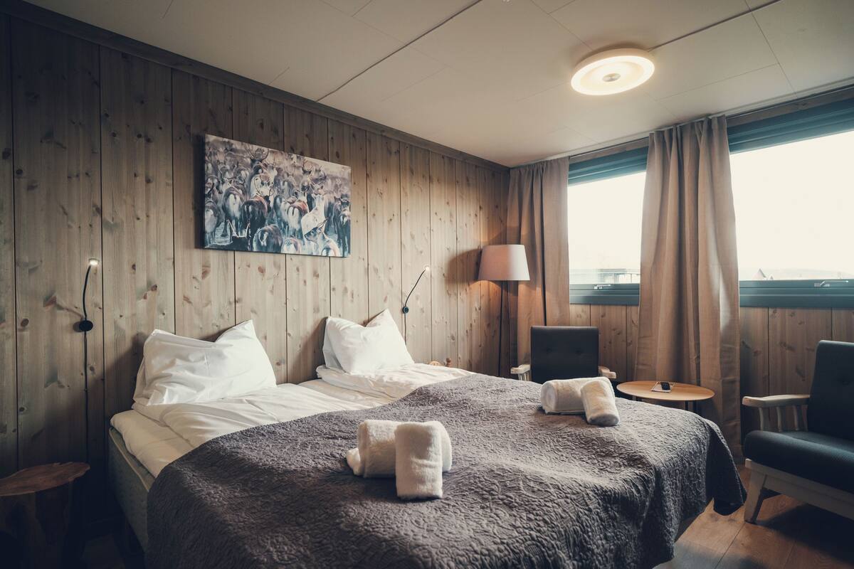 Habitación de hotel con pared de madera, una cama grande con ropa blanca, un cuadro, un escritorio con lámpara y vista al mar a través de una ventana.