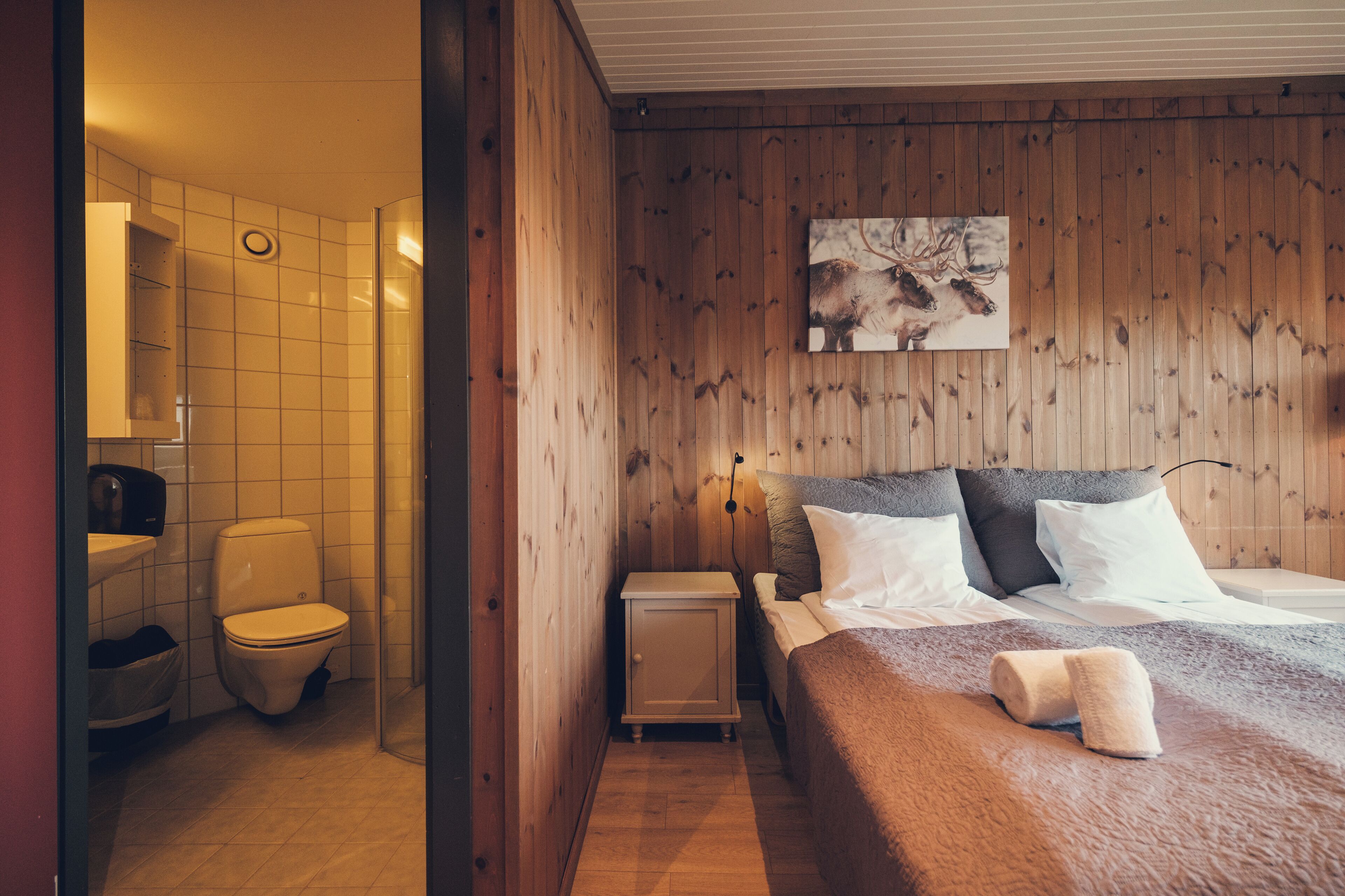 Una habitación con paneles de madera, una cama, una mesita de noche, un inodoro blanco y una zona de ducha.