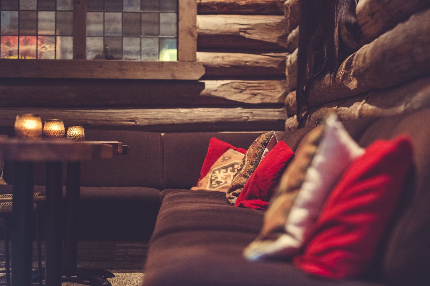 A cozy log cabin interior with a wooden log wall, a dark brown sofa with red and patterned cushions, and a small table with candles.