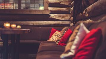 A cozy log cabin interior with a wooden log wall, a dark brown sofa with red and patterned cushions, and a small table with candles.