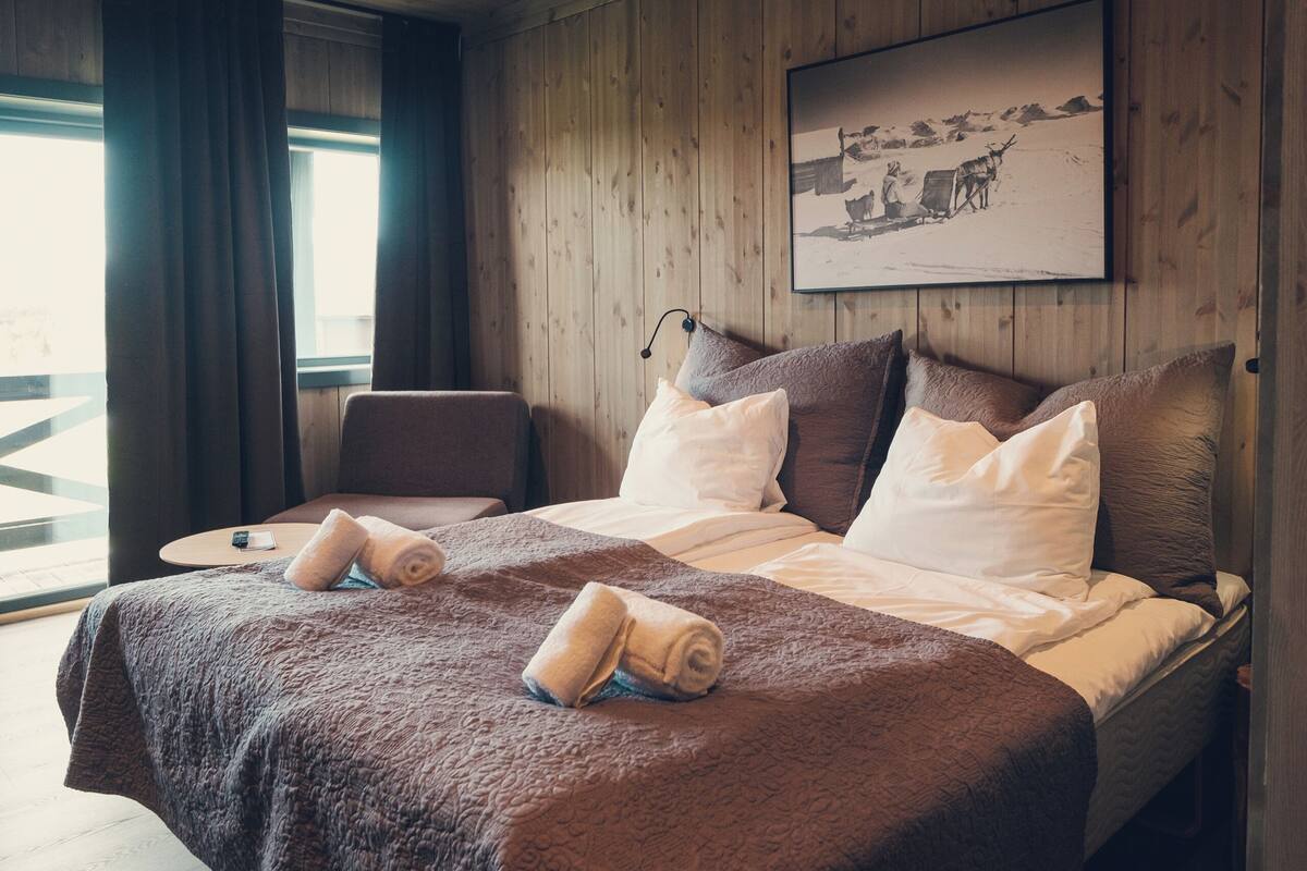 Un dormitorio con paneles de madera, una cama grande, dos almohadas, una mesita de noche y una ventana con cortinas.