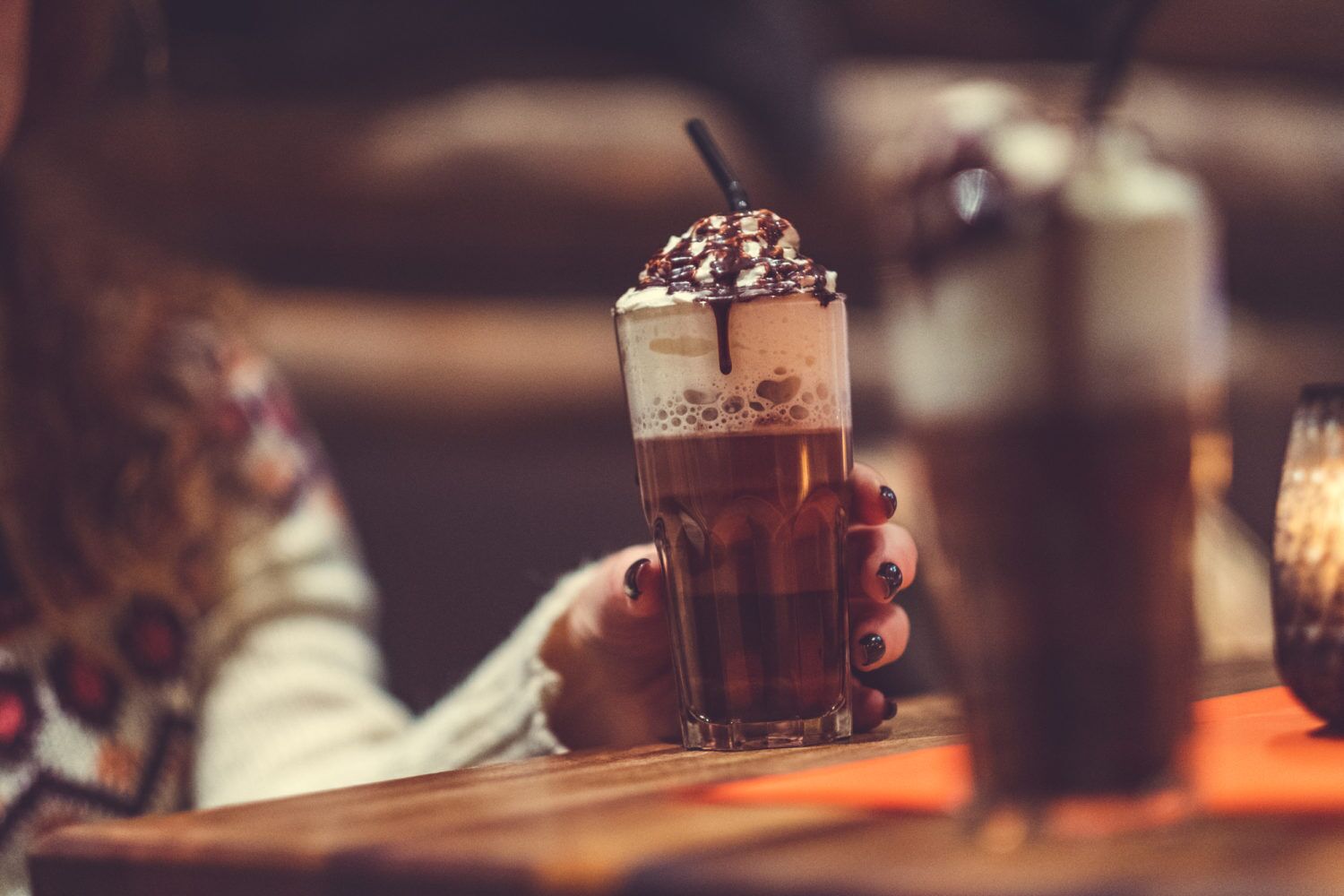 A person holding a glass of iced coffee with whipped cream and chocolate drizzle, with another glass in the background.