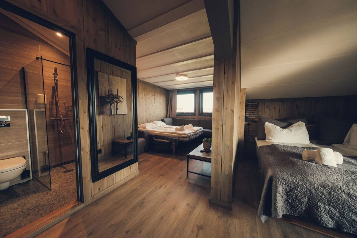 Una habitación tipo cabaña de madera con una cama, una mesita, un banco y una zona de ducha con mampara de vidrio.