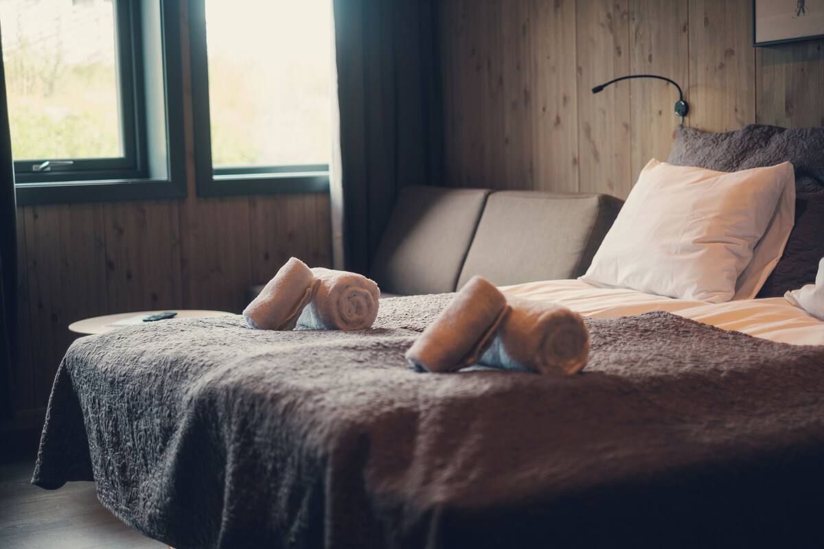 Una cama bien hecha con toallas enrolladas, una mesita de noche y una ventana con cortinas.