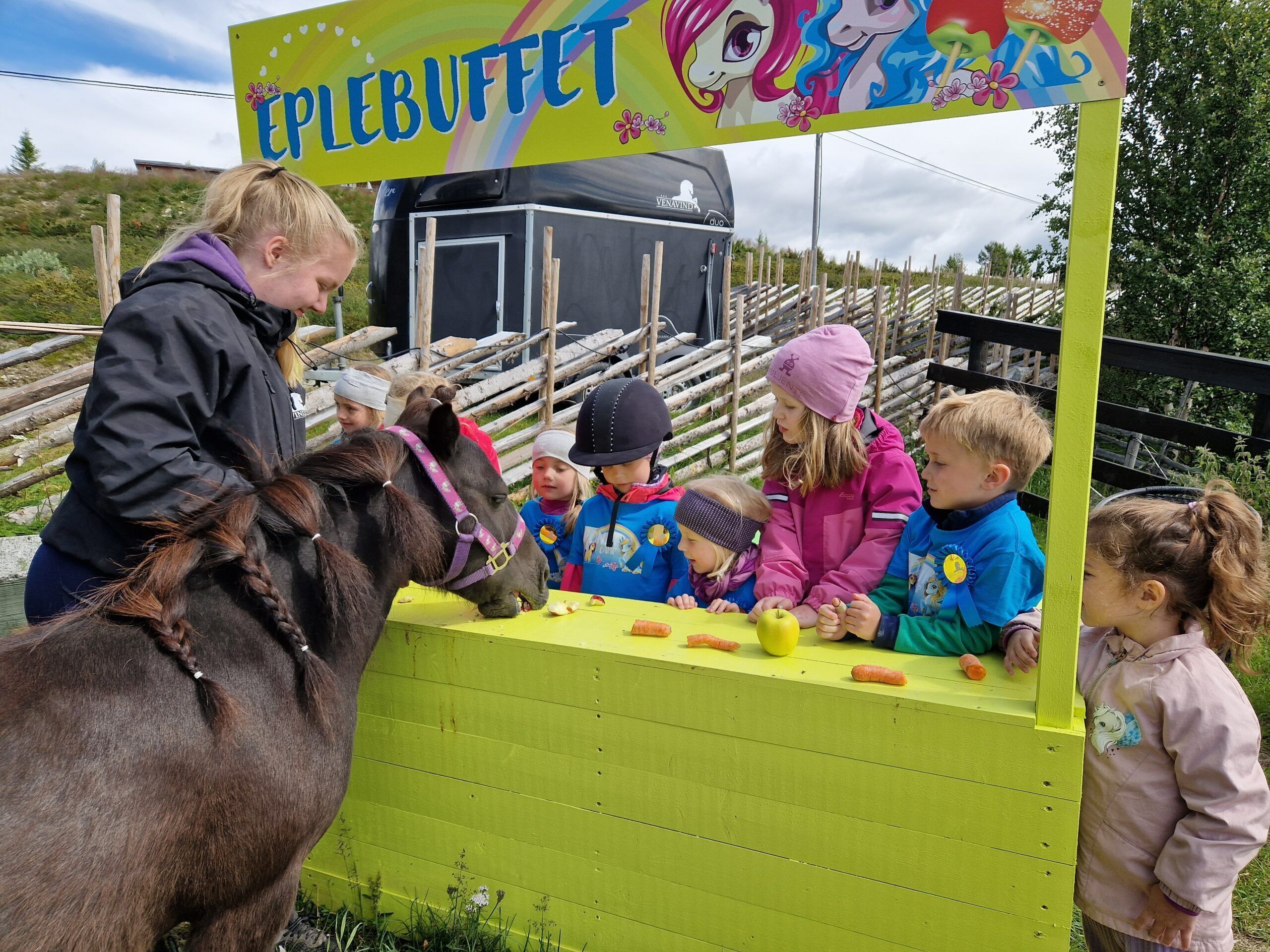 En plats där man kan åka ponny, med ett grönt staket, en skylt med texten ”ÄPPELBUFFÉ” och ett svart fordon i bakgrunden.