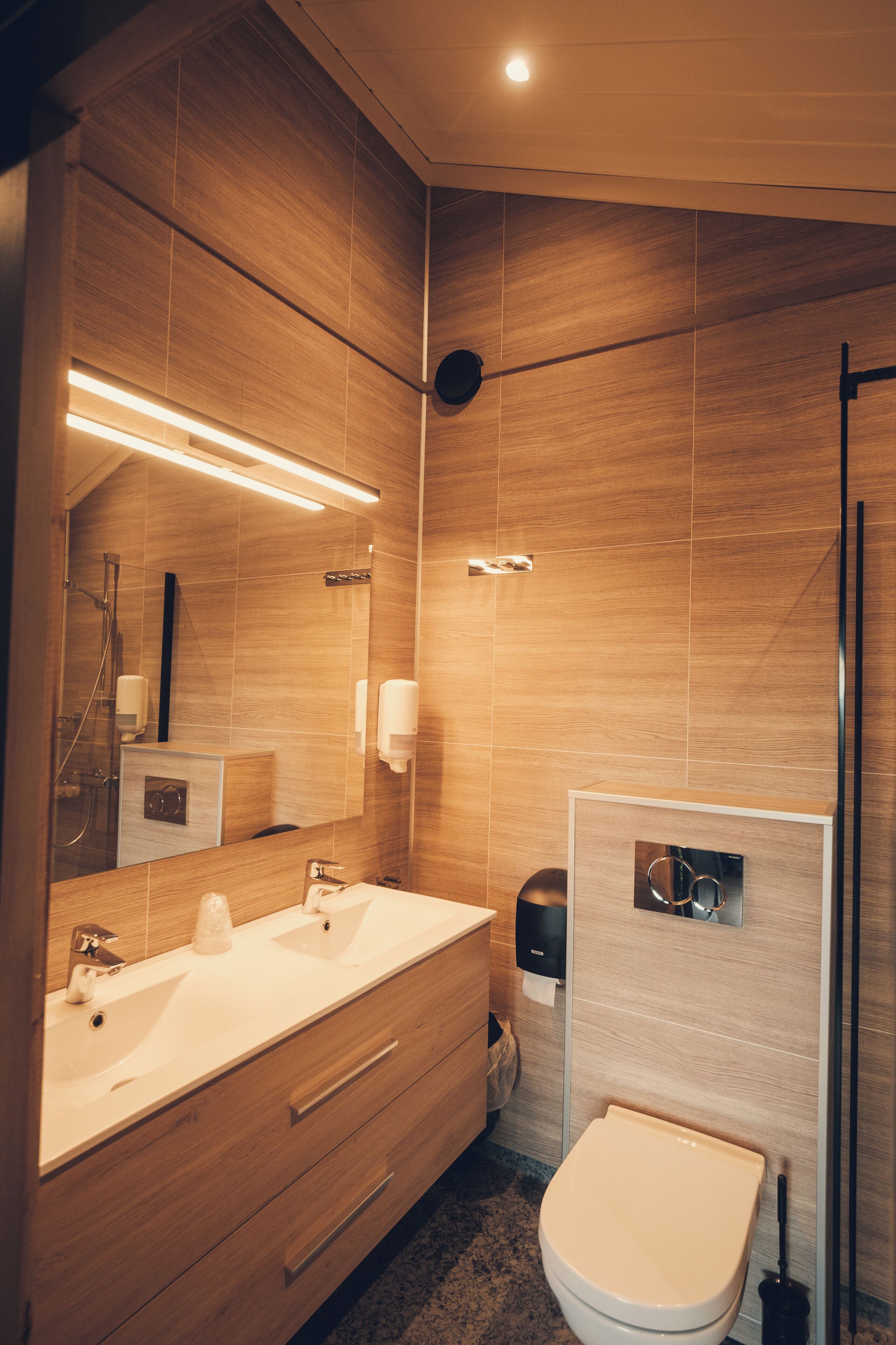 Une salle de bain moderne avec un lambris en bois, un grand miroir, un meuble-lavabo à deux vasques, des toilettes et un espace douche.
