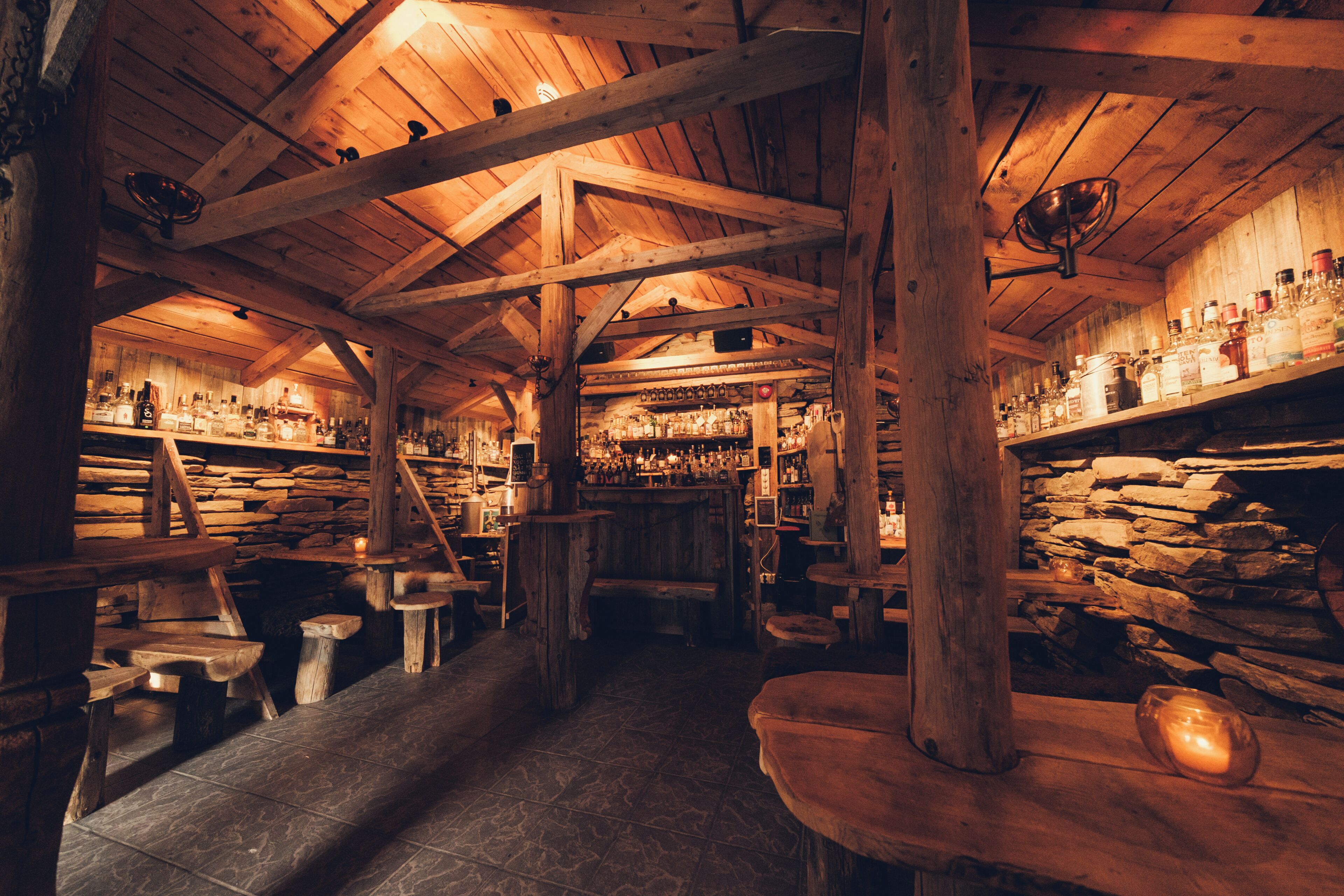 A rustic interior with wooden beams, stone walls, and a bar area with shelves of bottles.