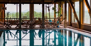 An indoor swimming pool with wooden deck chairs and tables, large windows with a view of greenery.