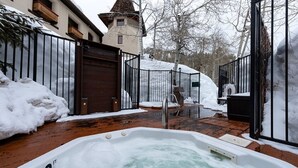 Un jacuzzi en un área exterior nevada con cubierta de madera, barandilla de metal y una puerta de madera grande.