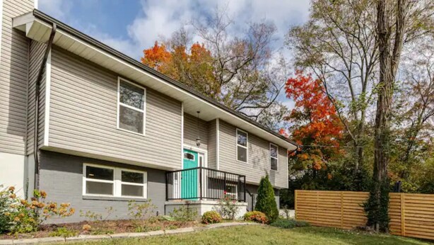 A two-story residential building with a front porch, a green door, and a small front yard with a wooden fence and trees.