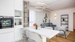Eine moderne Küche mit integriertem Backofen, ein Essbereich mit weißem Tisch und grauen Stühlen sowie ein graues Sofa im Wohnbereich.