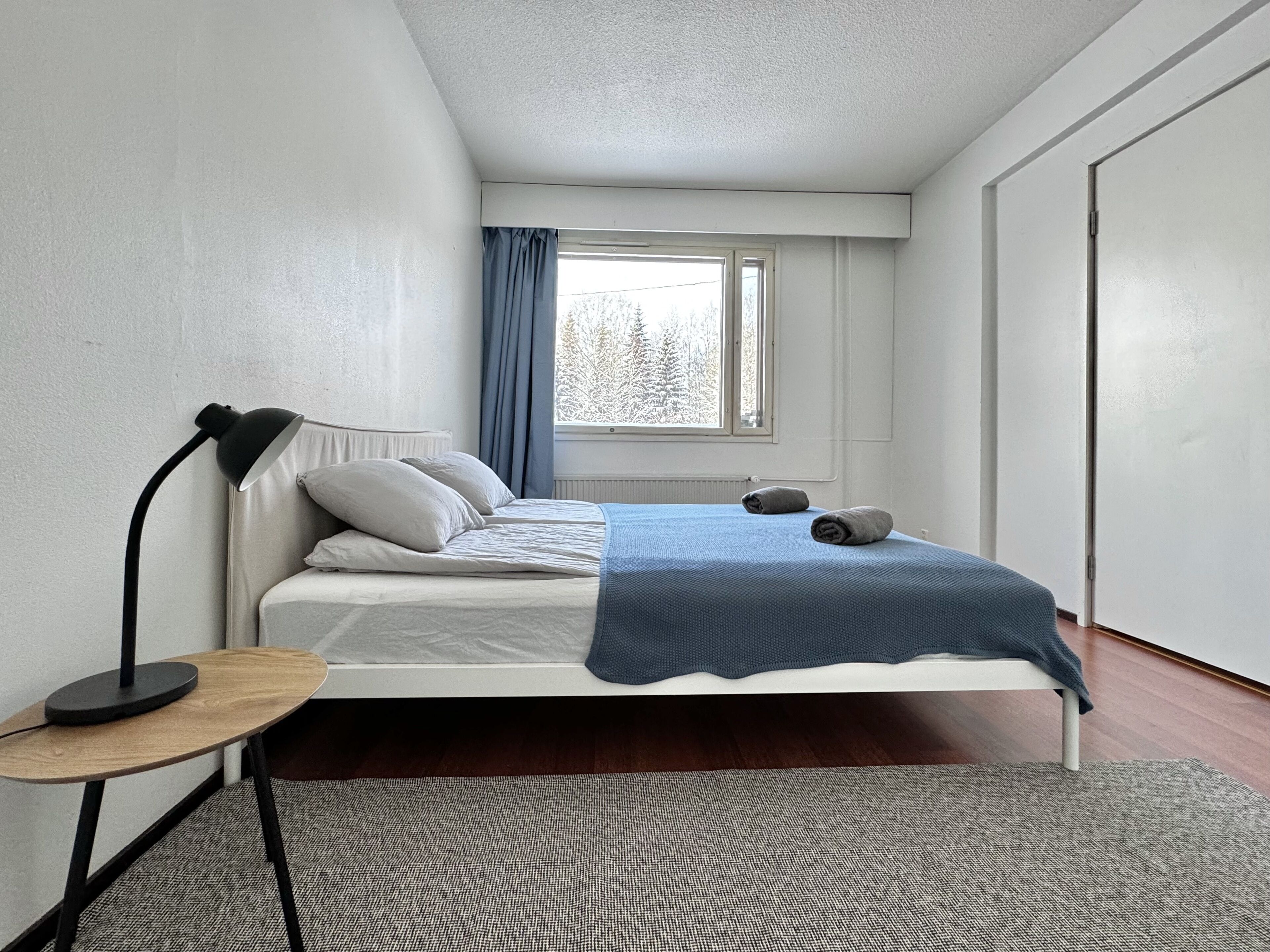 A bedroom with a single bed, a bedside table with a lamp, a window with curtains, and a view of trees outside.