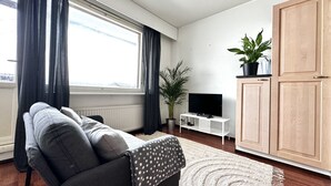 Un salon moderne avec un canapé gris, un téléviseur posé sur un meuble blanc, une grande fenêtre avec des rideaux, une plante en pot et des meubles en bois.