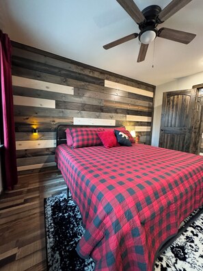 A bedroom with a wooden slat wall, a ceiling fan, a bed with a red and black checkered bedspread, and a black and white rug. - Hit the trail from the property! Game room, fire pit and 4 king suites (Muse)