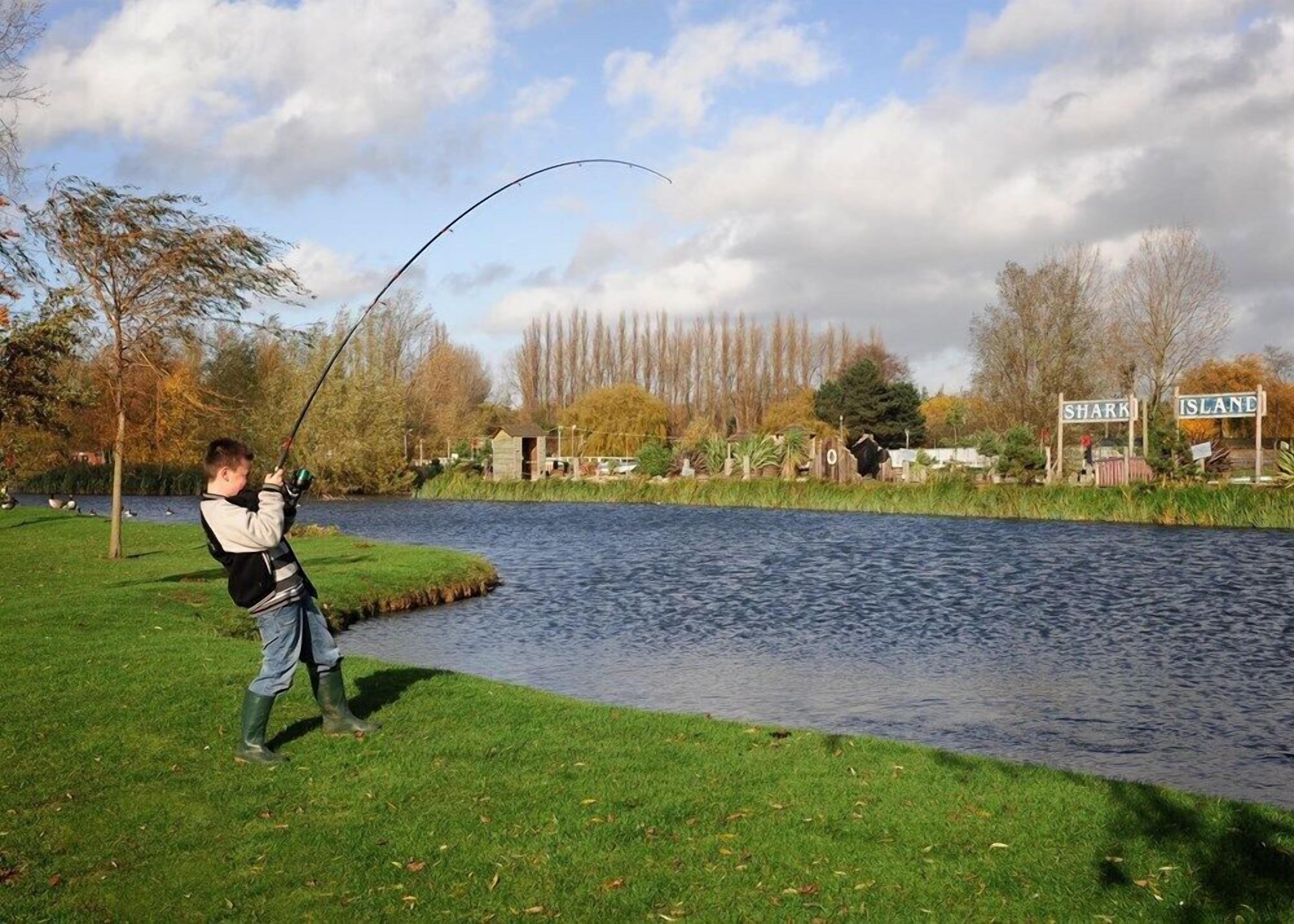 A person fishing by a calm river with grassy banks and trees in the background.