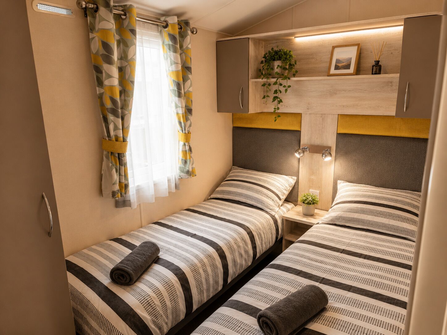 Ein gemütliches Schlafzimmer mit zwei Betten, einem Fenster mit Vorhängen, einem Nachttisch mit Lampe und einem Regal mit Pflanze und Bild.
