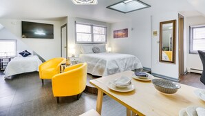 Una habitación de hotel moderna con una cama, dos sillas amarillas, una mesa de comedor con platos, un televisor y un ventanal grande.