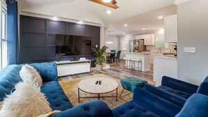 A modern living room with a large flat-screen TV, blue sofas, a round coffee table, and a ceiling fan. - Charlotte Pet Friendly 3BR Chateau • Hot Tub • Mini Golf • Fire Pit • SouthPark (Charlotte)