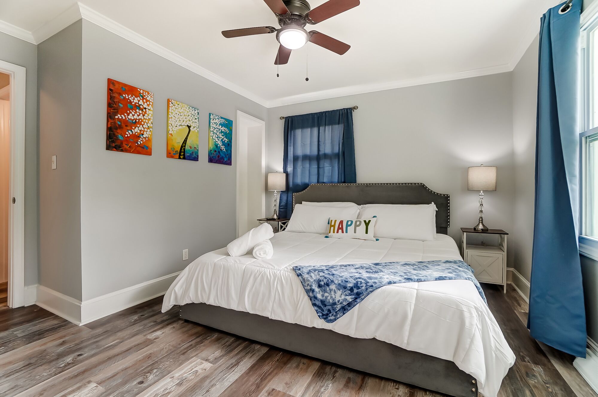 A bedroom with a bed, a ceiling fan, artwork on the wall, and a window with curtains.