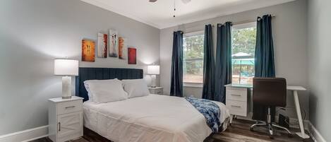 A bedroom with a bed, bedside tables, a desk, a chair, a ceiling fan, and artwork on the wall.