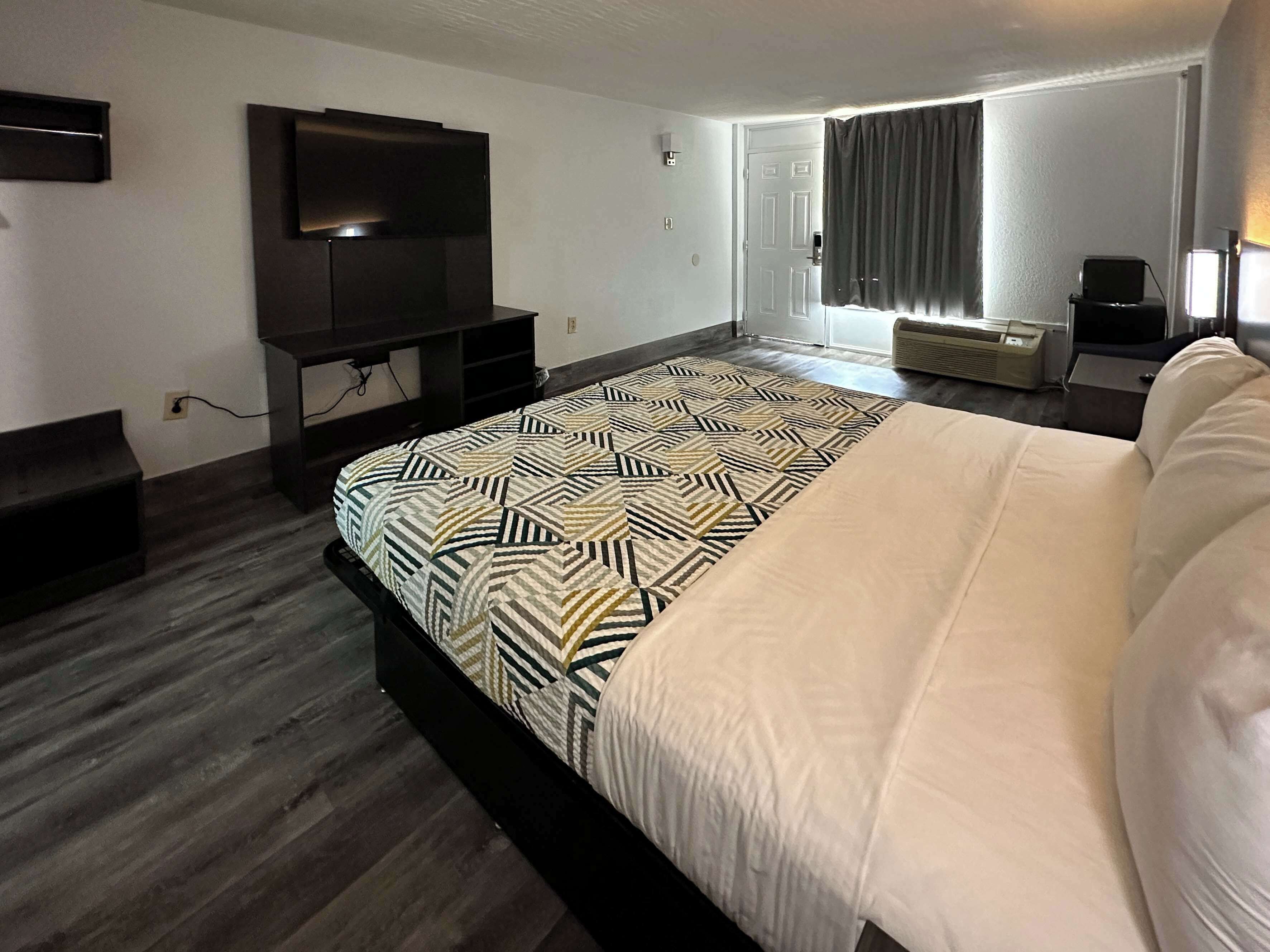 A hotel room with a large bed, a flat-screen TV mounted on the wall, a heater, and a window with curtains.