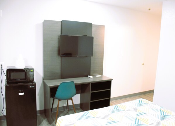 A compact hotel room with a bed, a desk with a chair, a microwave, and a television mounted on the wall.