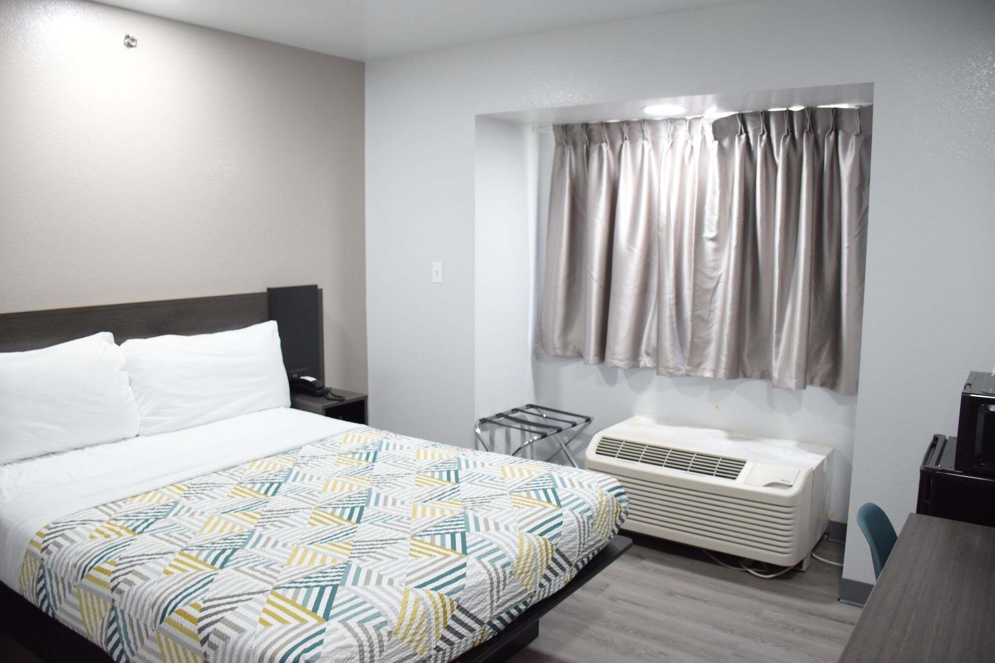 Une chambre d’hôtel comprenant un lit, une table de chevet, un bureau, une chaise, une fenêtre avec des rideaux et un appareil de climatisation.