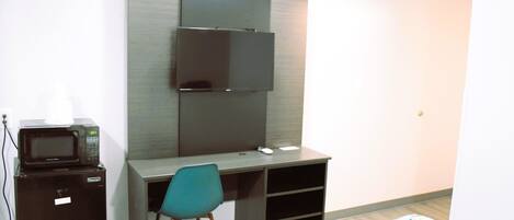 A compact hotel room with a single bed, a desk with a chair, a TV mounted on the wall, and a microwave on a small cabinet.