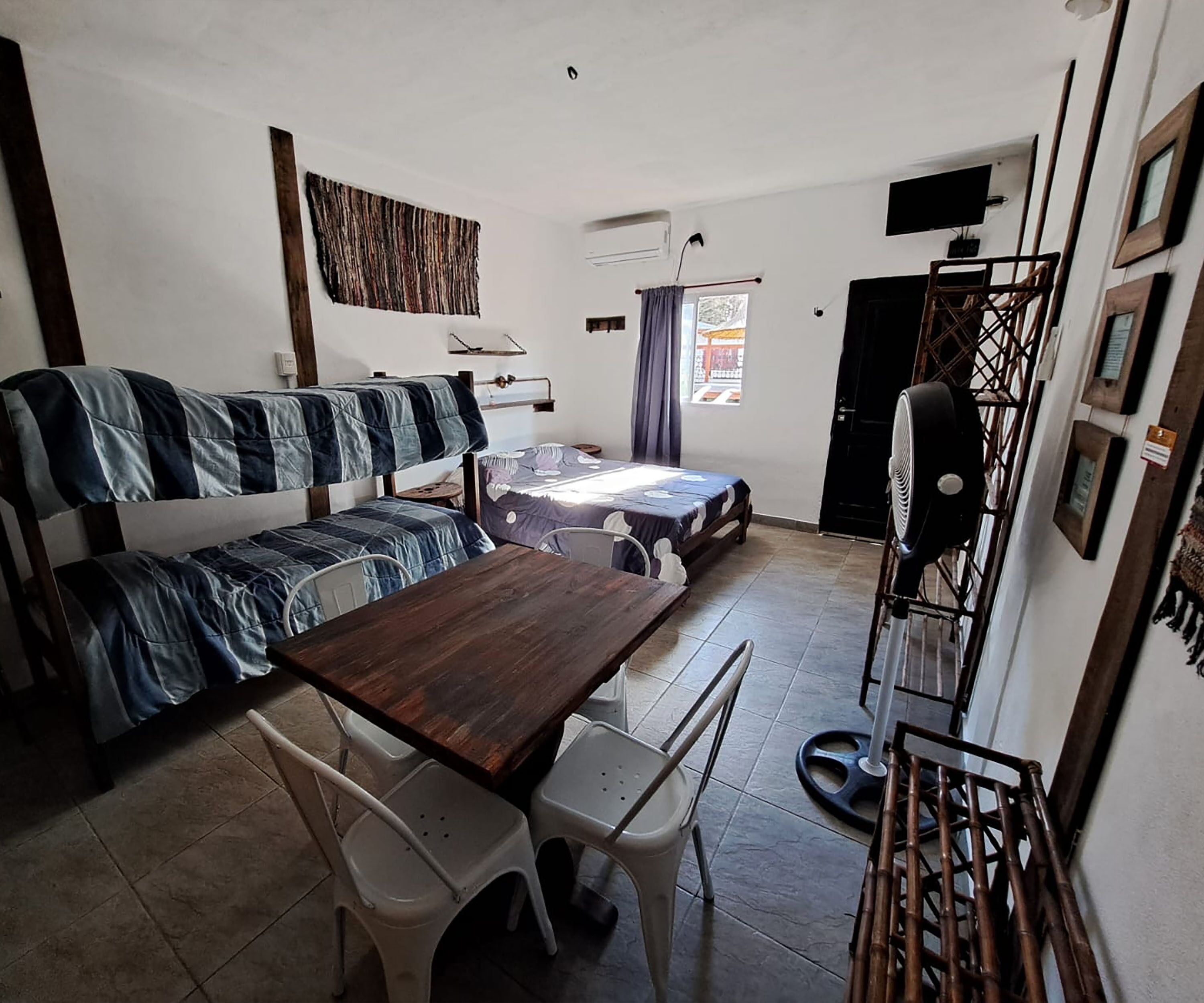 Une petite pièce avec des lits superposés, une table à manger avec des chaises, un ventilateur et un téléviseur fixé au mur.