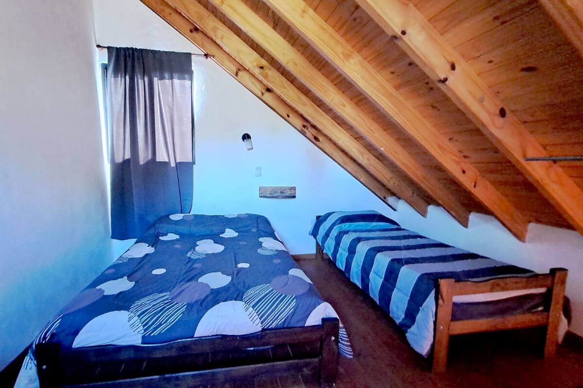 Une chambre avec un plafond en pente, un lit recouvert d’une couvre-lit à motifs, une table de chevet et une fenêtre avec des rideaux.