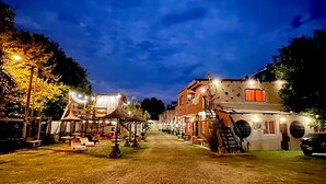 A rustic building with a staircase leading to the upper floor, surrounded by trees and a well-lit outdoor area with tables and chairs.