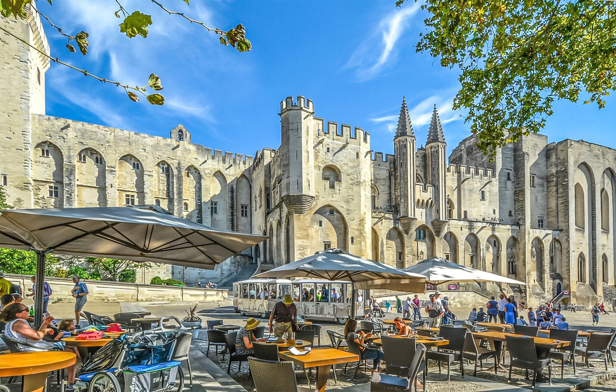 An outdoor café with tables and umbrellas, set against a historic building with arches and towers.