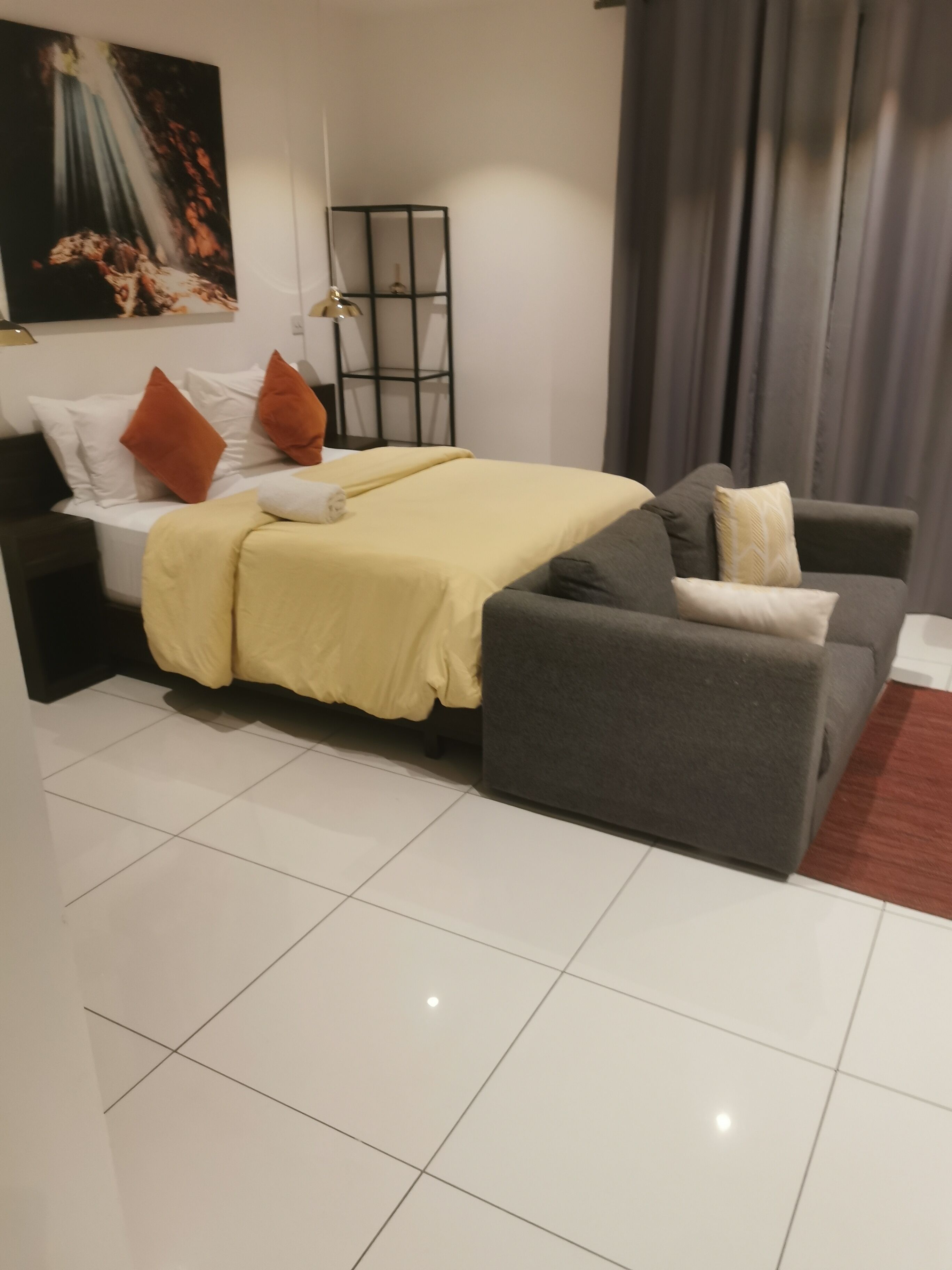 A modern hotel room with a bed, a grey sofa, a small table, and a painting on the wall.