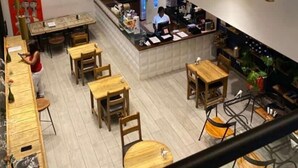 A multi-level restaurant with wooden tables and chairs, a counter with a staff member, and exercise equipment on the upper level.
