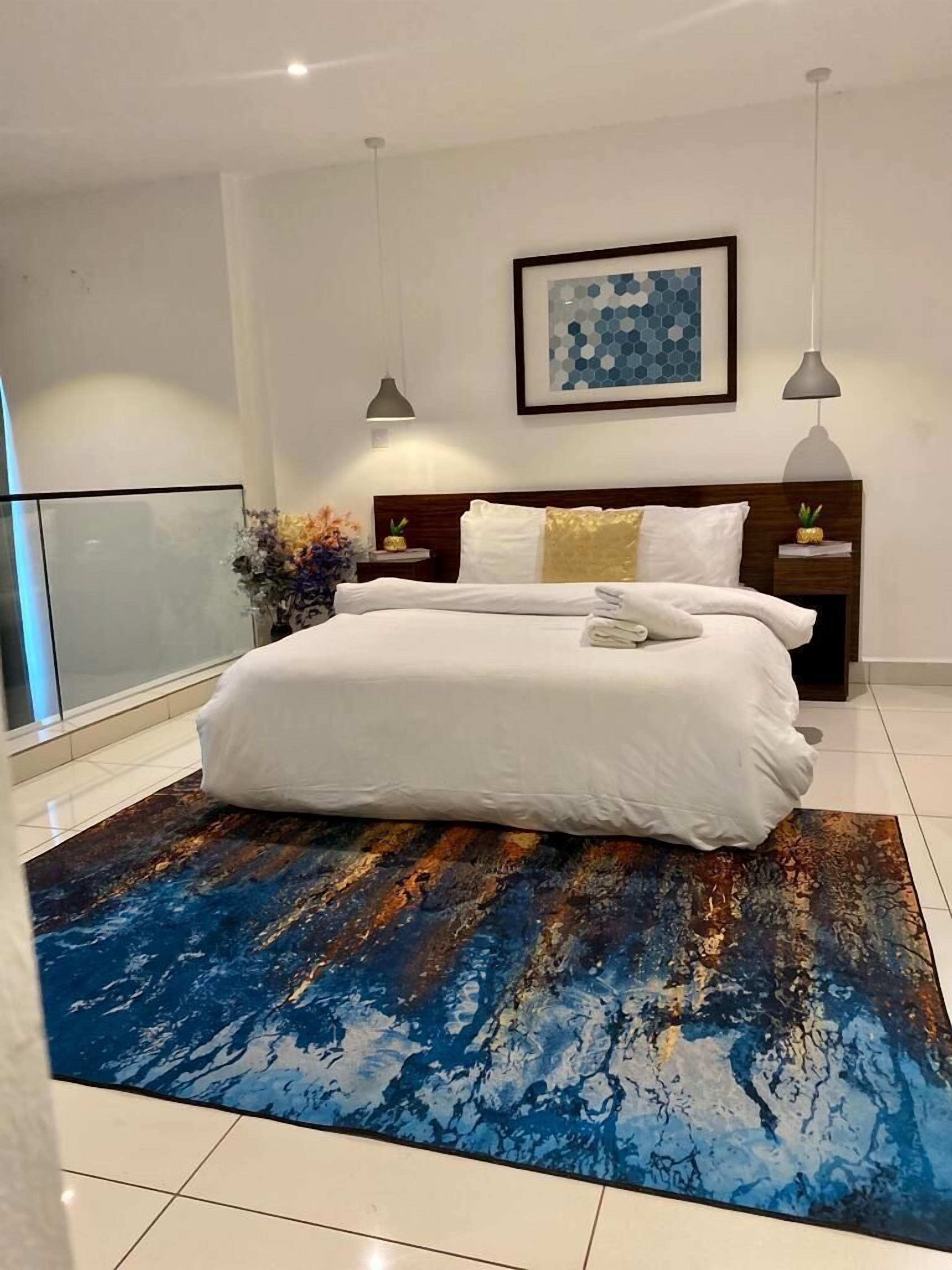 A modern bedroom with a large bed, a blue abstract rug, a framed artwork on the wall, and a nightstand with a plant.