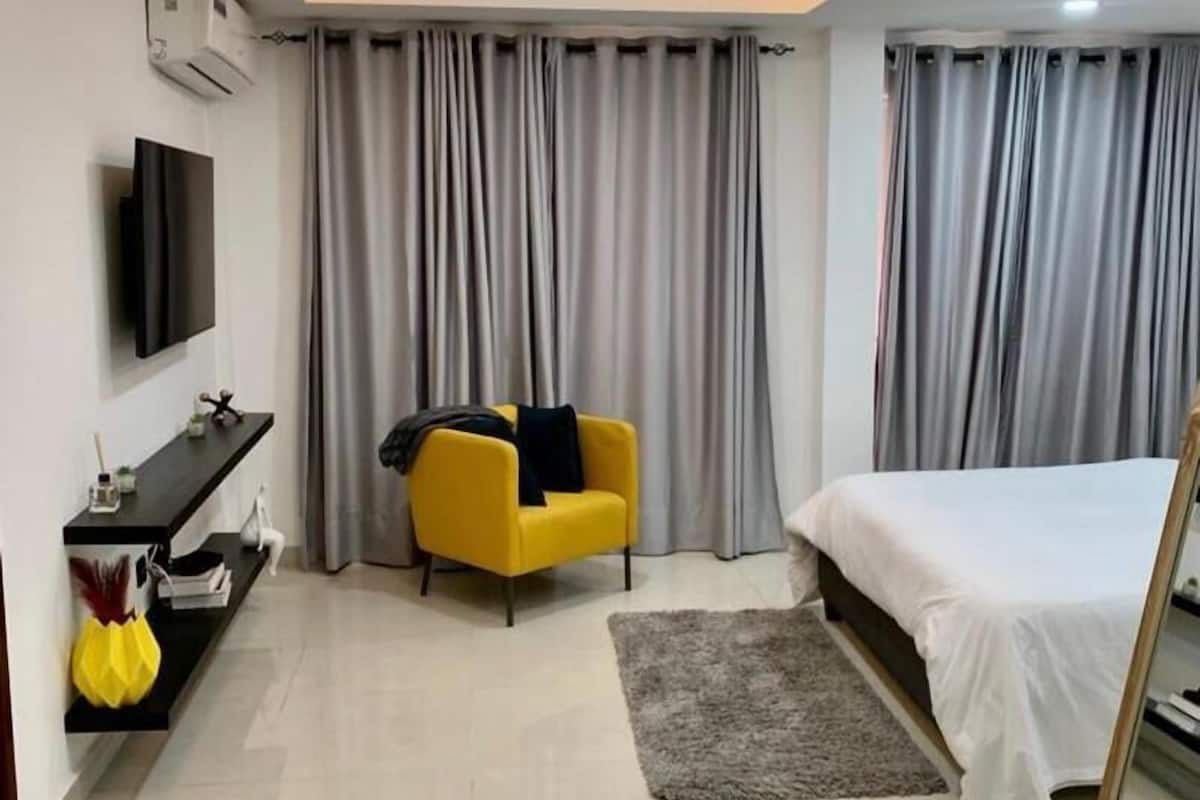Habitación de hotel moderna con una cama, un sillón amarillo, un escritorio con televisor de pantalla plana y cortinas grises.