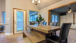 Un comedor con una mesa de madera, sillas y un banco. Hay ventanas con persianas, una araña y una puerta.