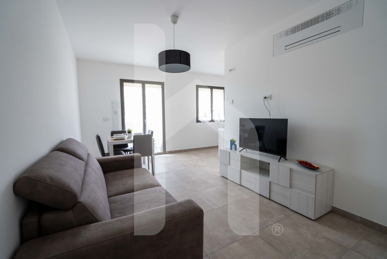 Una sala moderna con un sofá marrón, un televisor de pantalla plana sobre un mueble blanco y un comedor al fondo.