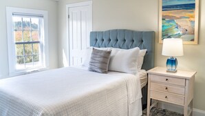 A bedroom with a large bed, a nightstand with a lamp, a window with a view of trees, and a painting on the wall. - Cozy second floor suite with kitchenette! (Hampton)