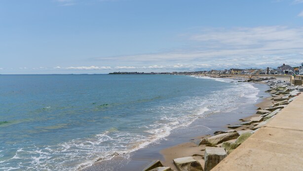 A coastal view with a sandy beach, a rocky breakwater, and a distant shoreline with buildings. - Gorgeous Oceanfront Suite! (Hampton)