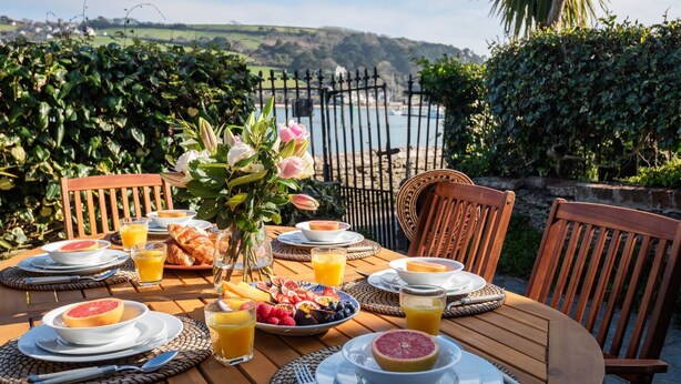 A table set for breakfast with plates of fruit, croissants, and orange juice, overlooking a scenic view of hills and a body of water. - Lifeboat Cottage -  a cottage that sleeps 2 guests  in 1 bedroom (Salcombe)