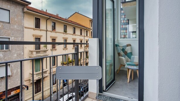Ein Balkon mit Blick auf benachbarte Gebäude, eine Glastür, die zu einem Innenraum mit einer Couch und einem kleinen Tisch führt.