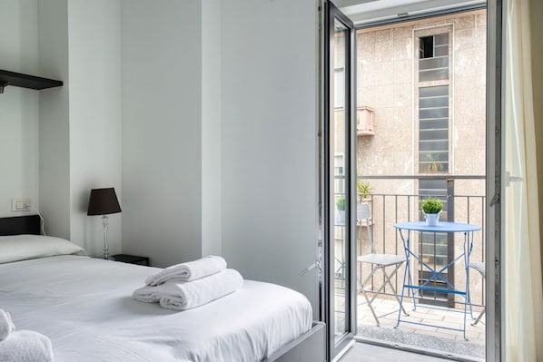 Une chambre d’hôtel avec un lit, une table de chevet, une lampe, un balcon avec une table et des chaises, et une plante en pot.