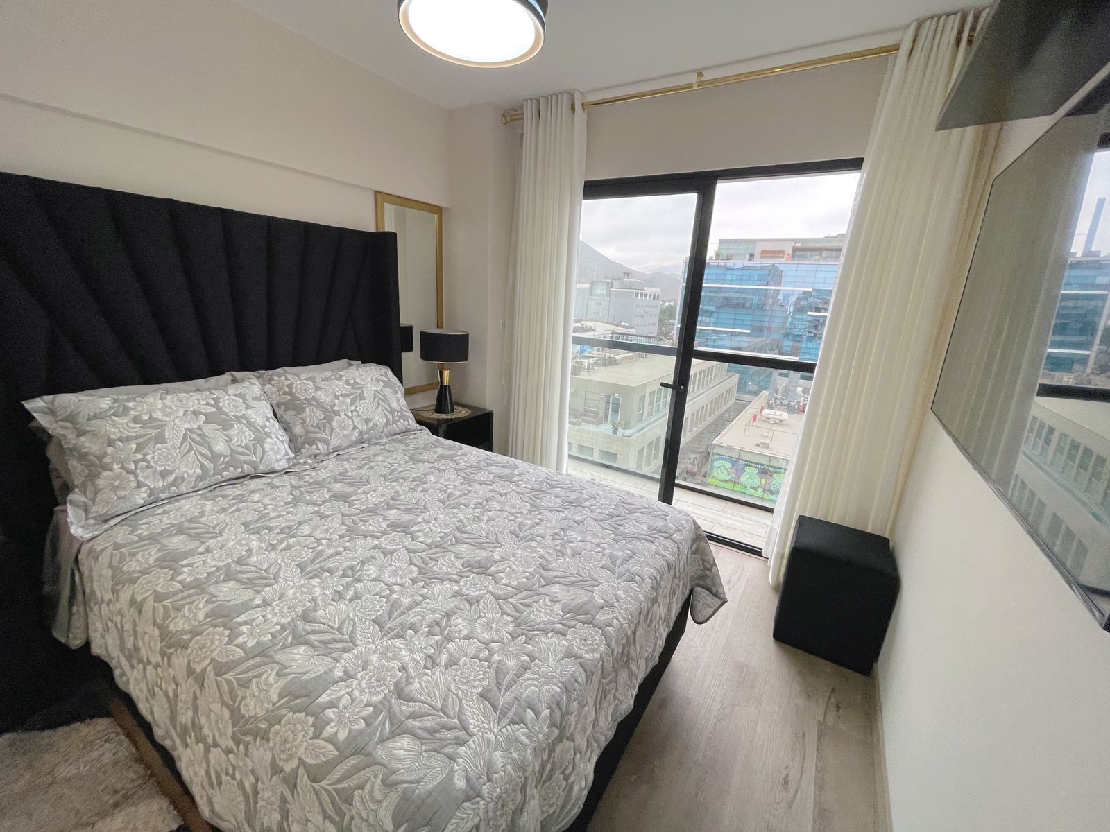A modern bedroom with a large bed, a nightstand, a lamp, and a view of the city through a large window.