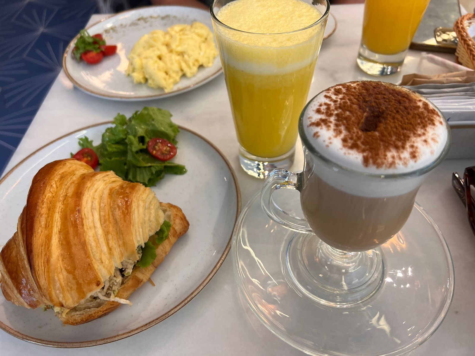 A breakfast setting with a croissant sandwich, scrambled eggs, a glass of orange juice, and a cup of coffee with whipped cream and cinnamon.