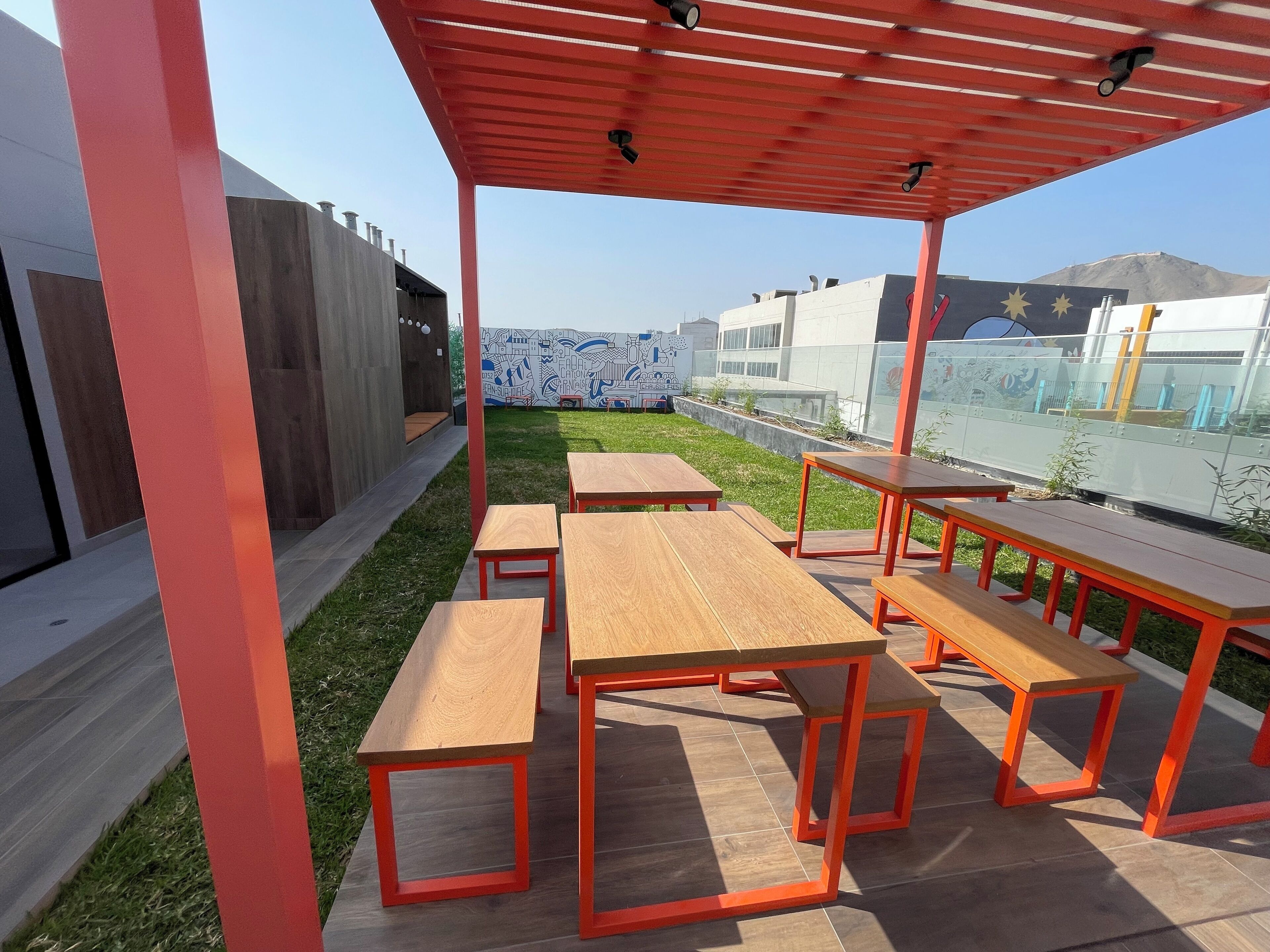 Un área de estar al aire libre con mesas y bancos de madera bajo un toldo rojo, rodeada por un césped y una pared con murales.