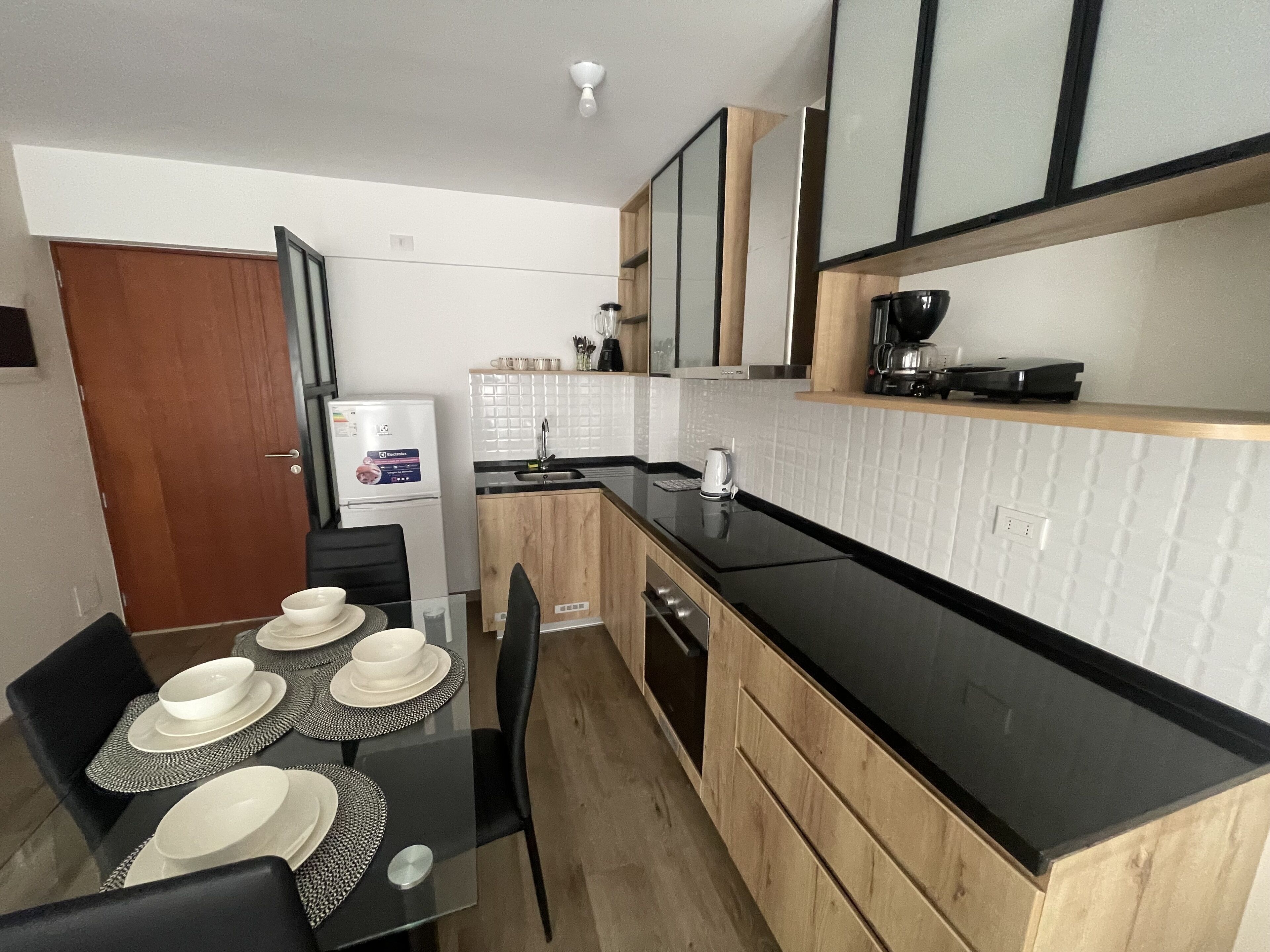 Une cuisine moderne avec des meubles en bois, un plan de travail noir et un coin repas doté d’une table en verre, préparée pour accueillir quatre personnes.
