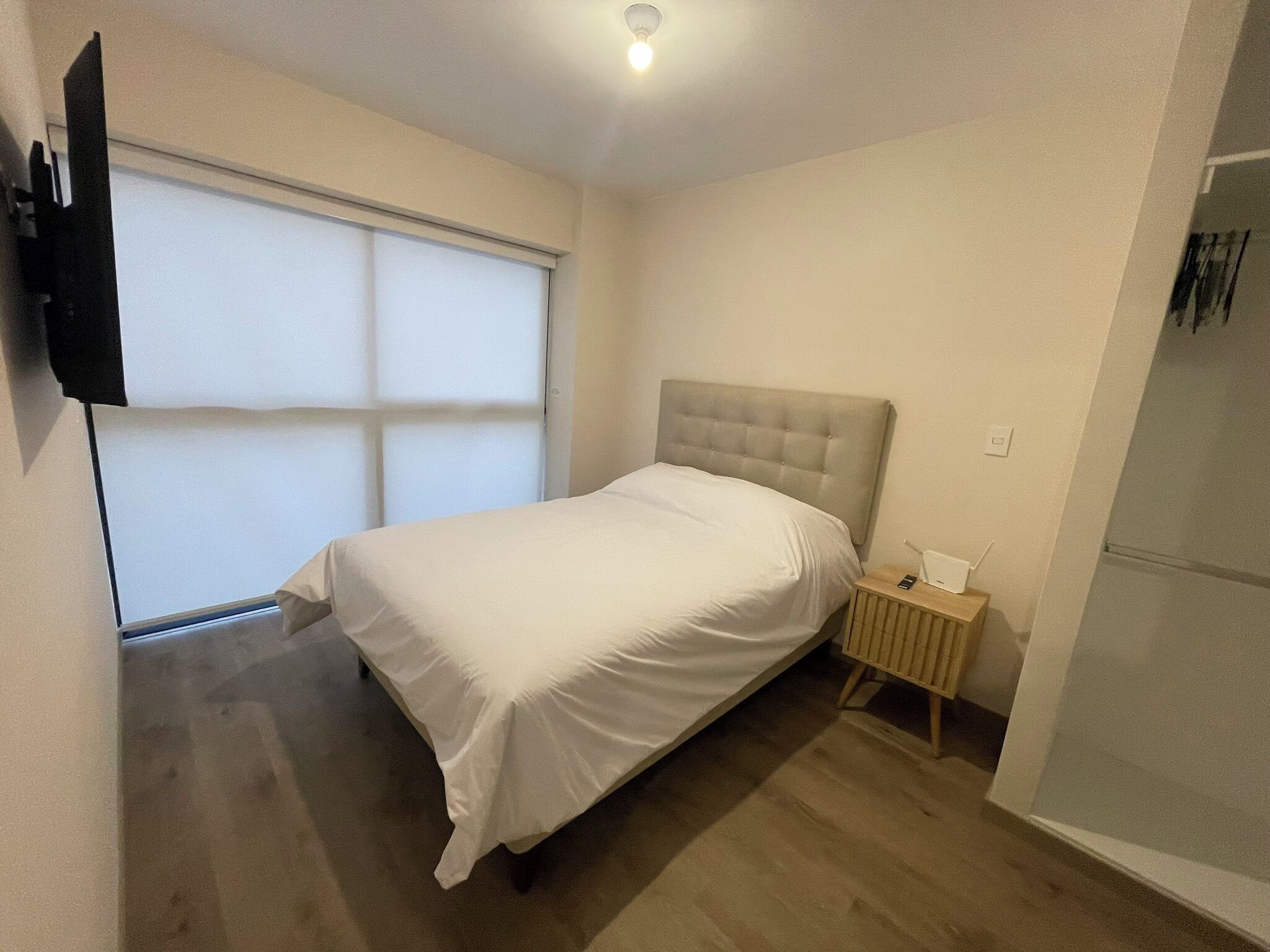 A bedroom with a bed, a bedside table, a TV mounted on the wall, and a window with blinds.