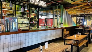 Un espace bar avec un mur recouvert de carreaux, des tables en bois et une variété de bouteilles et de verres exposés sur des étagères.